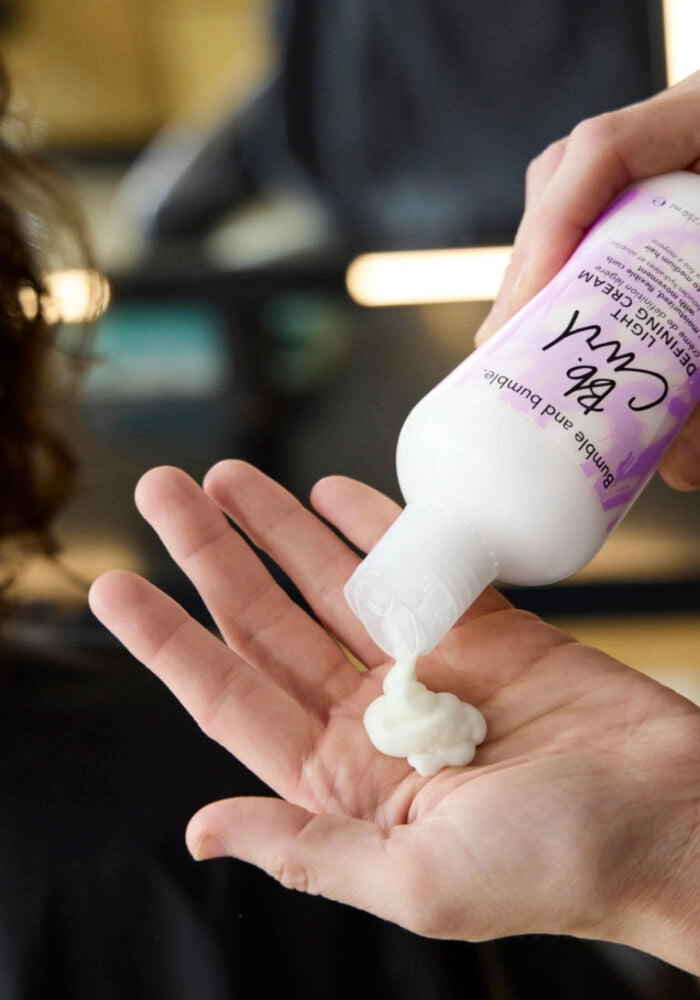 A close-up of a person pouring white hair-denying cream from a purple and white bottle into their palm, with the brand name 'The Curl' visible on the label.