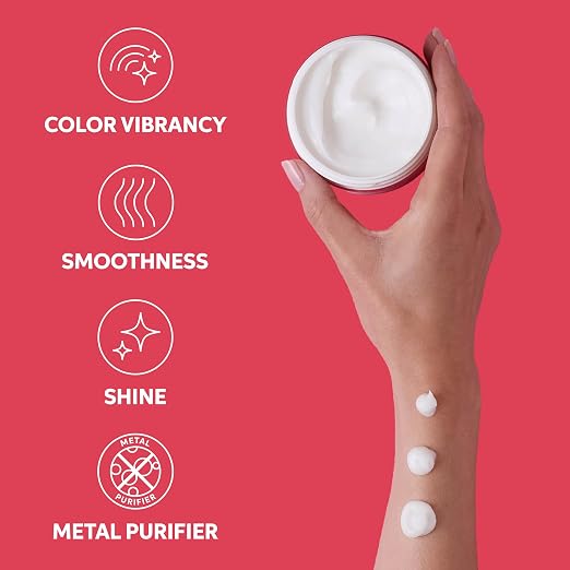 A hand holds an open white cosmetic jar against a bright pink background, with three dollops of white cream on the wrist. White text on the left lists benefits: color vibrancy, smoothness, shine, and metal purifier.